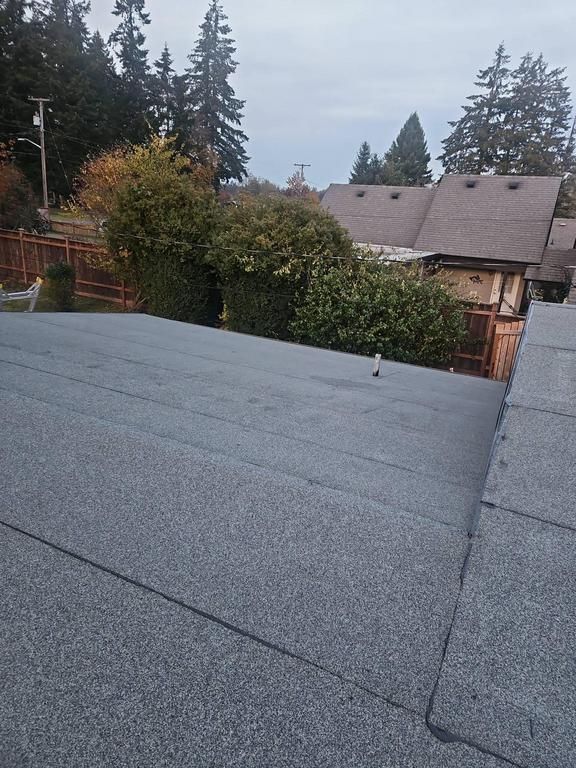 Flat roof with asphalt shingles, overlooking houses, trees, and cloudy sky.
