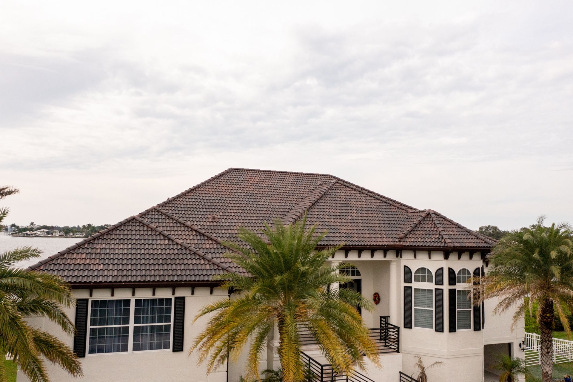 White house with dark tile roof and palm trees, overlooking water under cloudy sky.