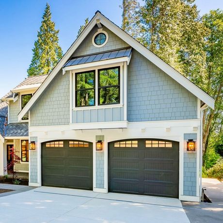 Gray two-story house with twin dark garage doors, white trim, and a steep gabled roof on a sunny day