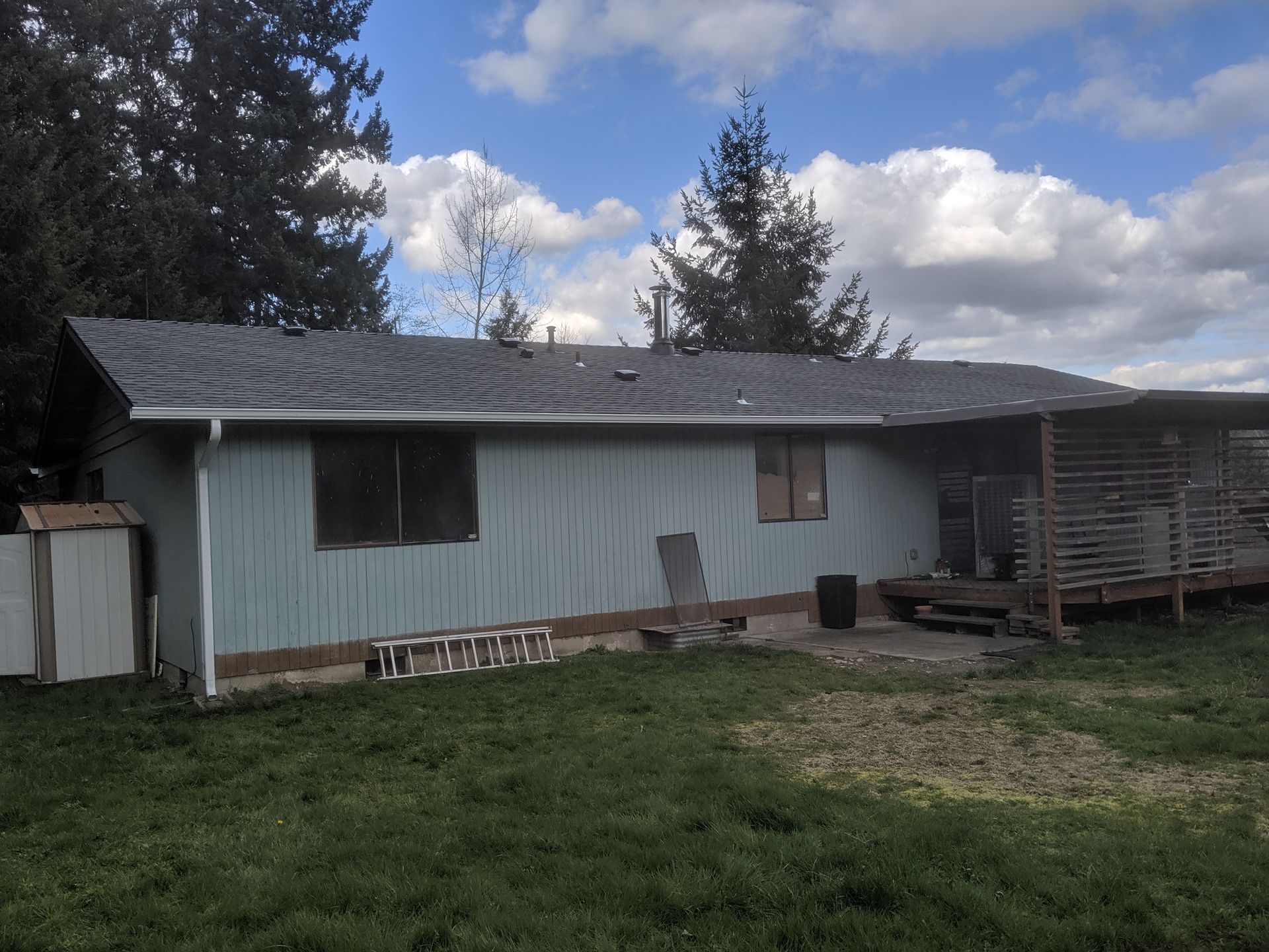 Back view of a house with light blue siding, damaged roof, and a covered patio, in a grassy area with trees.