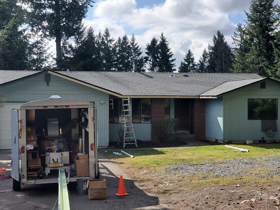 House with gray roof, construction trailer, ladder, and tools in yard; cloudy sky, green siding.
