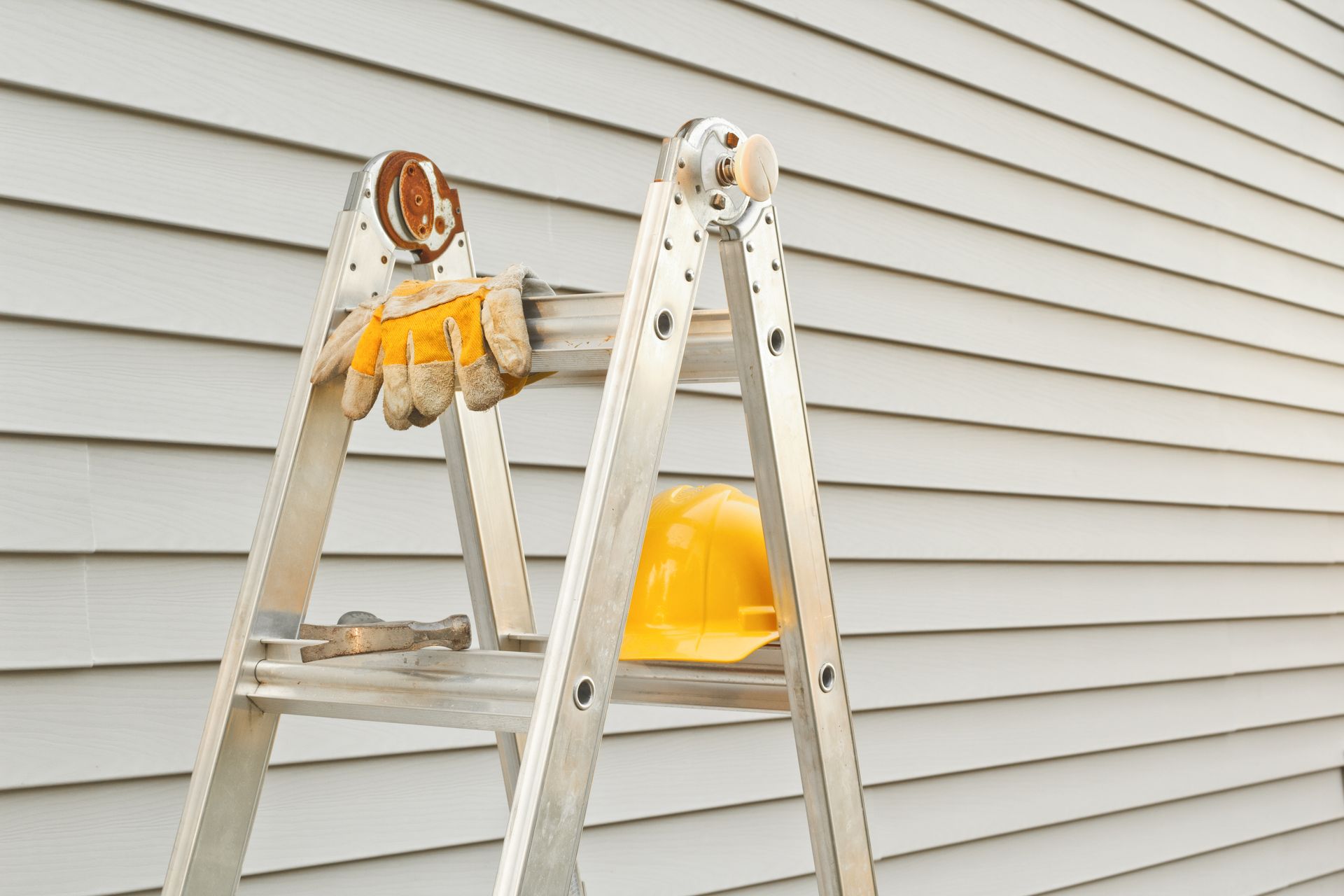 Aluminum ladder with gloves and yellow hard hat against a gray siding wall.