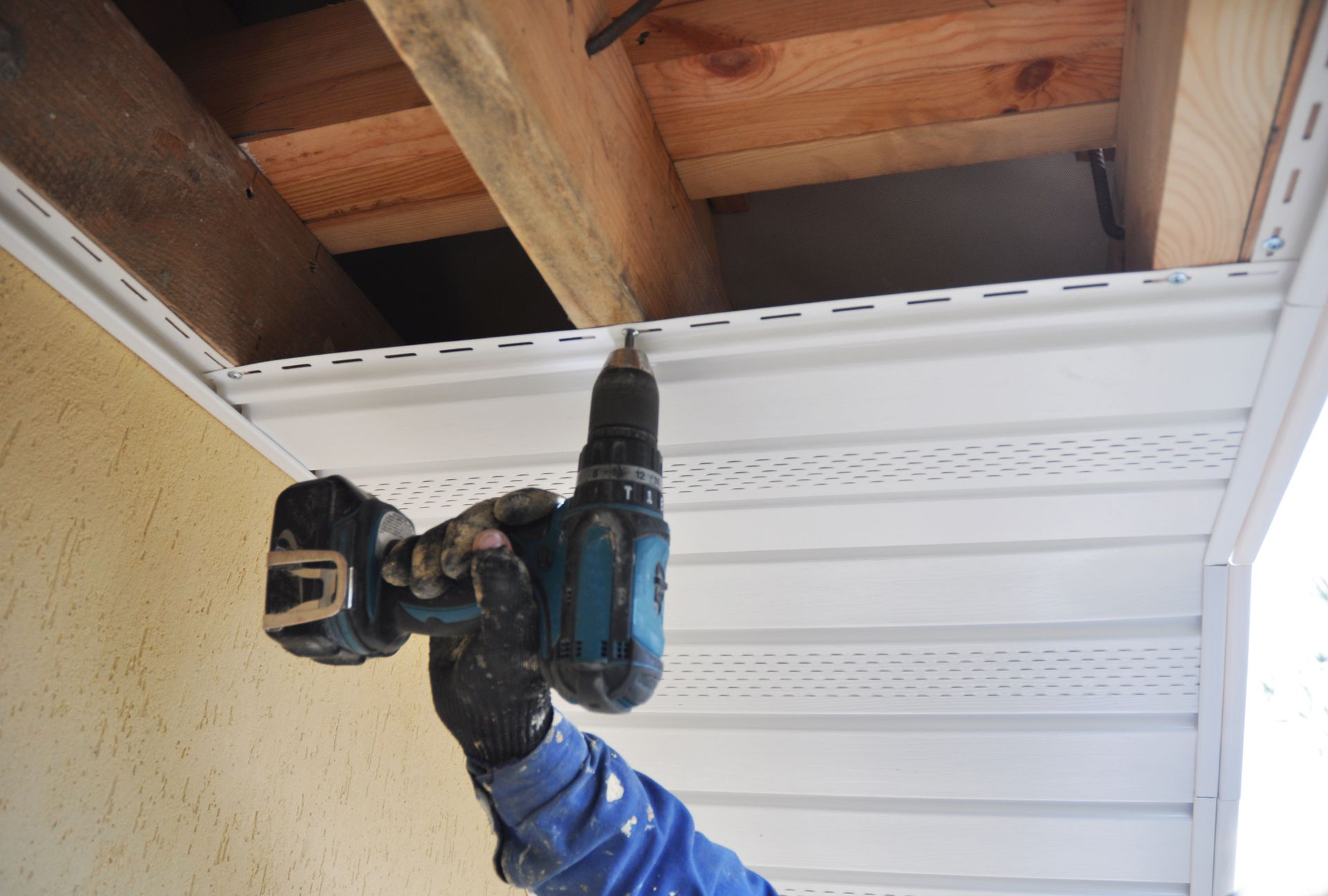 Person using a power drill to install white vinyl siding on a building's underside by Team Roofing NW in Kent, WA.