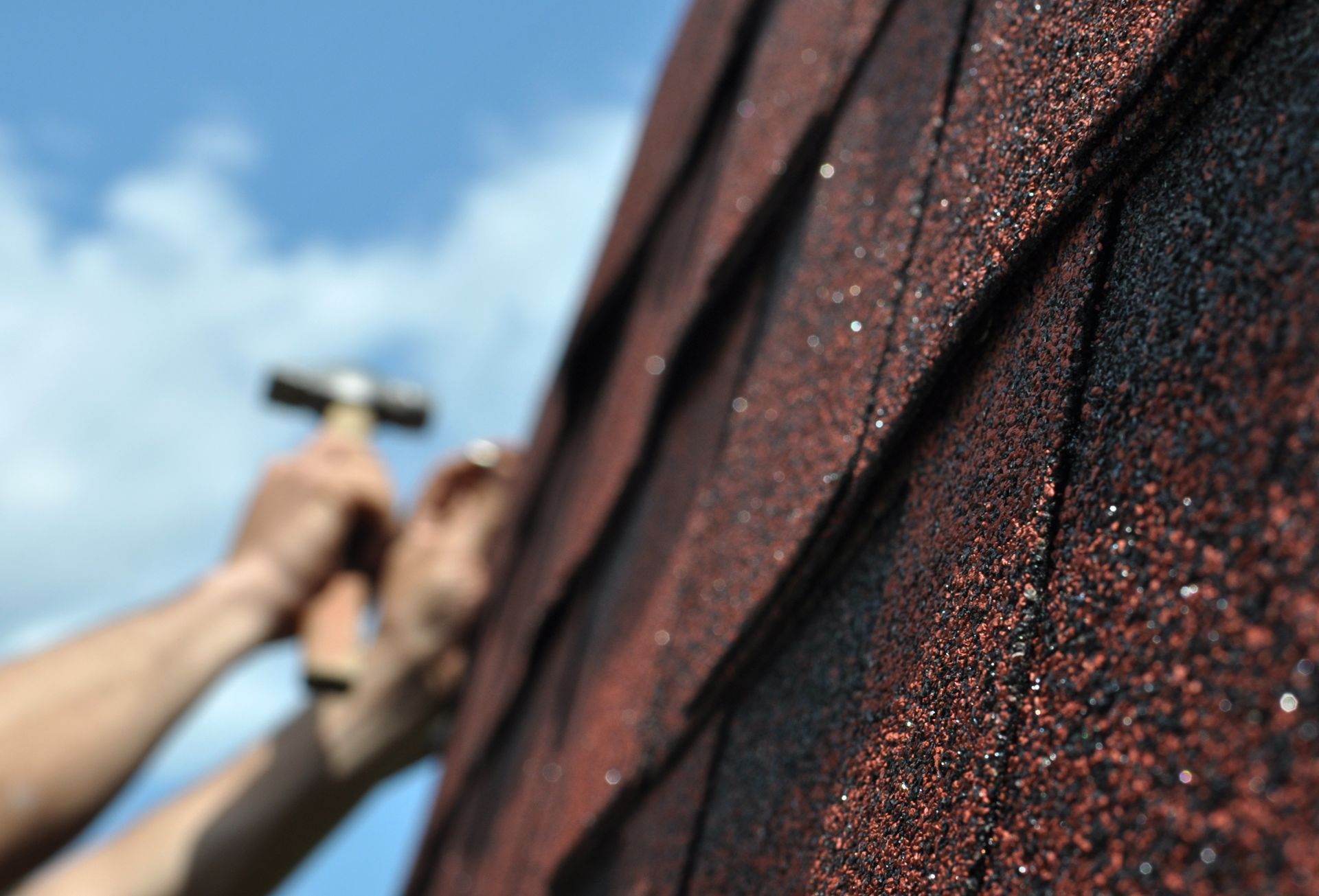Person hammering roofing shingle on a blue-sky day.