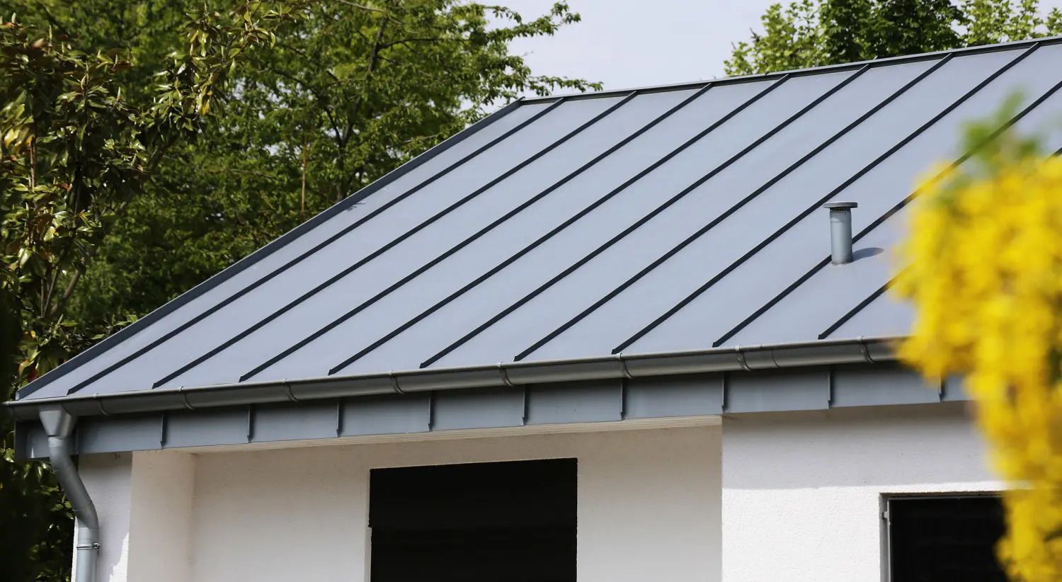 Grey metal roof on a white house with trees in the background.