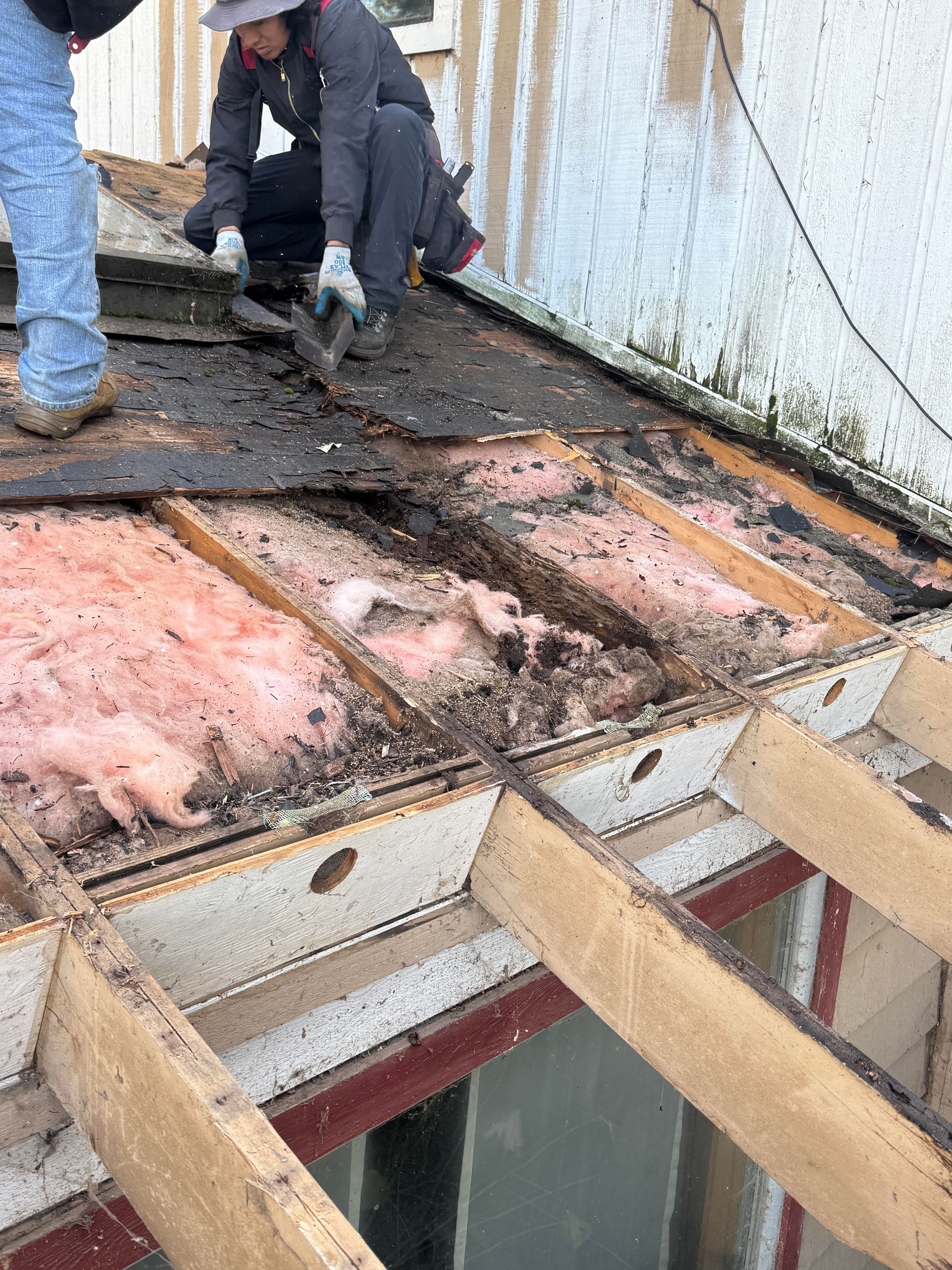 Workers removing roof materials, exposing insulation and wooden beams.