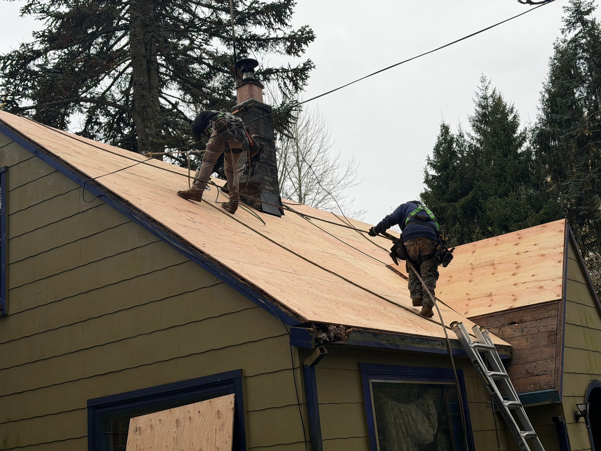 Two roofers working on a tan house roof, installing shingles near a chimney.