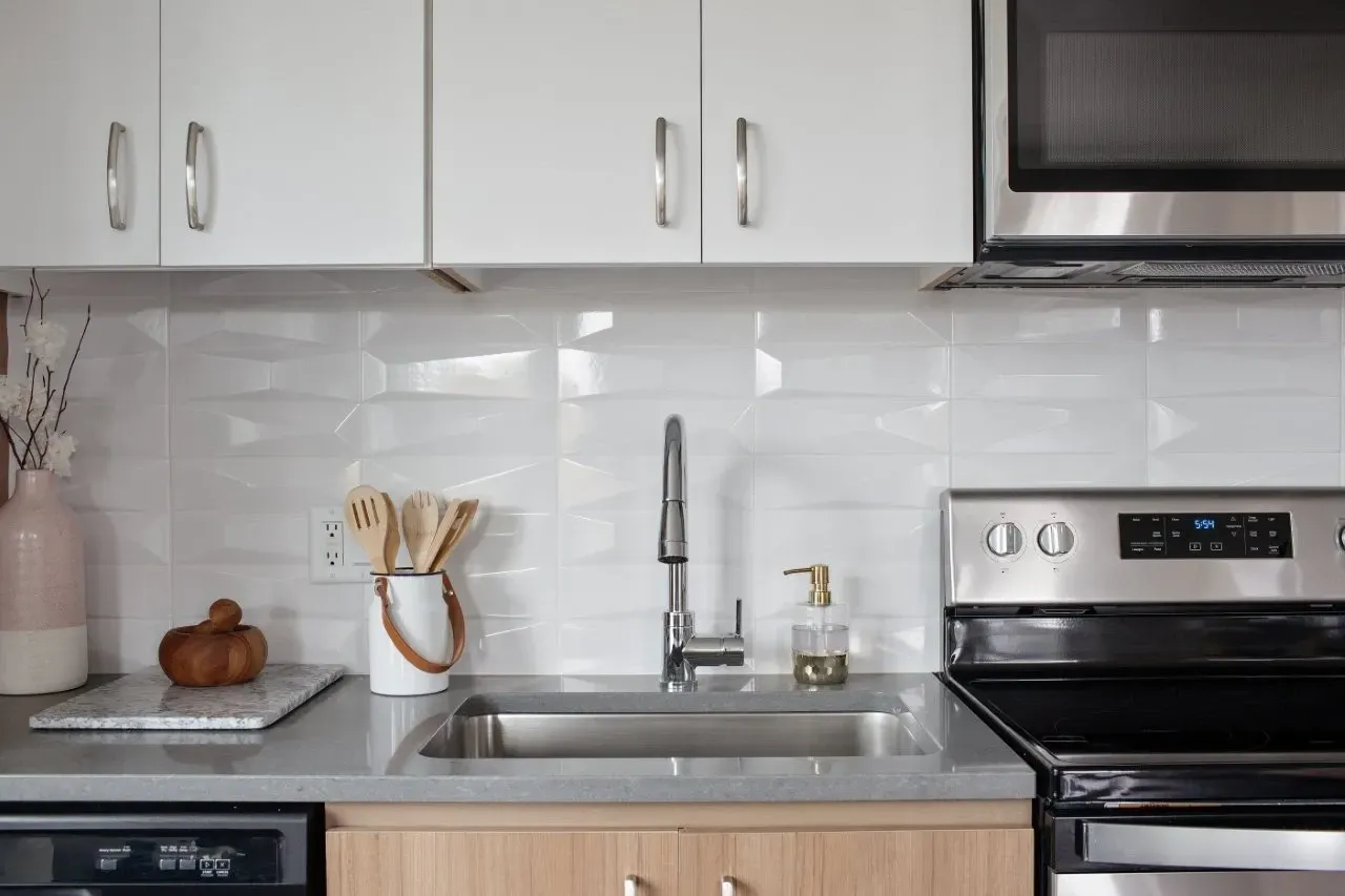 Kitchen sink with stainless steel faucet, white textured backsplash, and modern cabinetry.