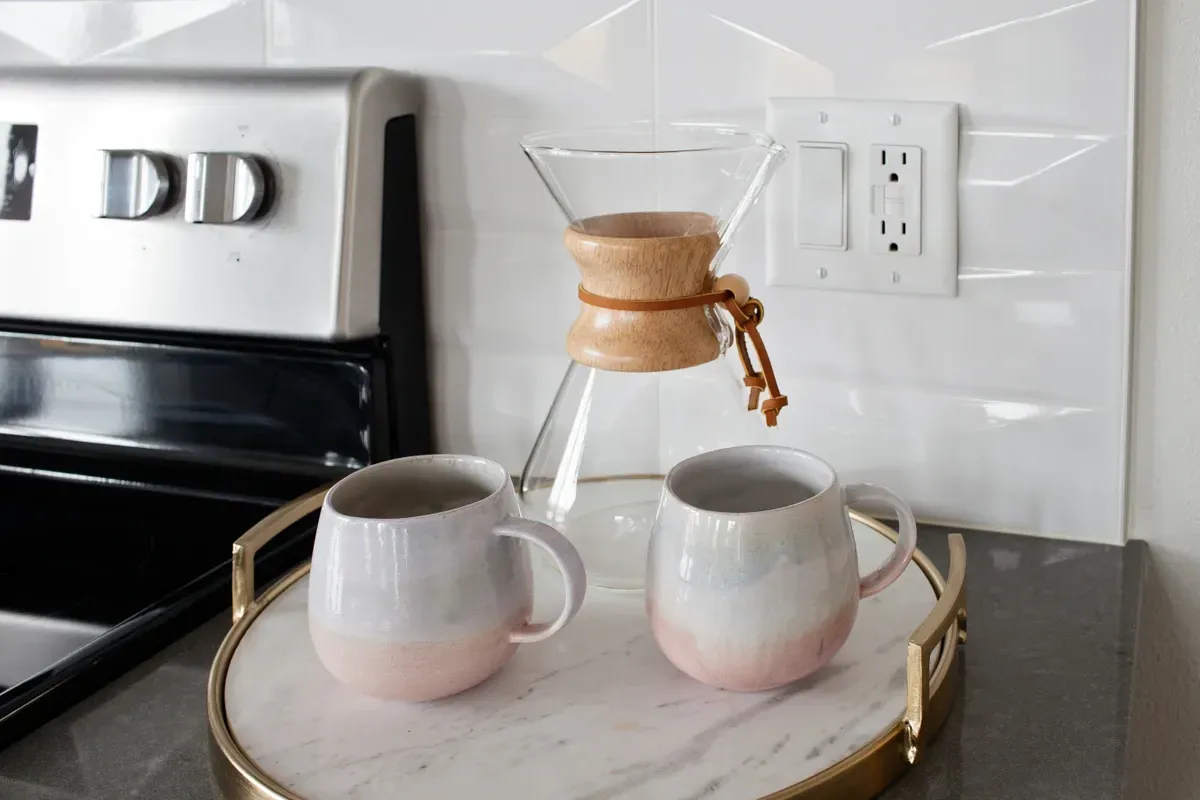 Two pink mugs on a marble tray with a coffee pot in the background.