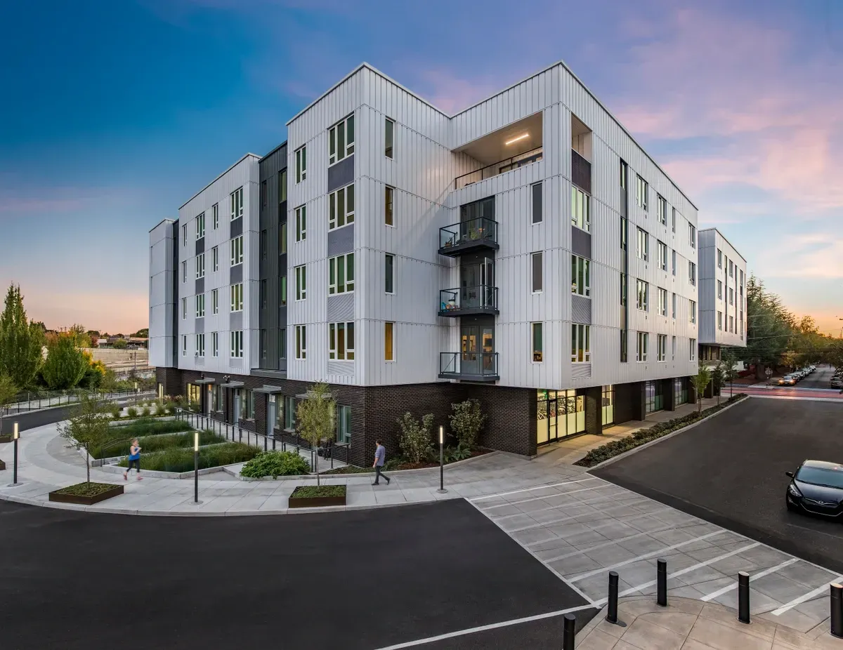 A modern building with a white facade and balconies is surrounded by a landscaped area and a road.