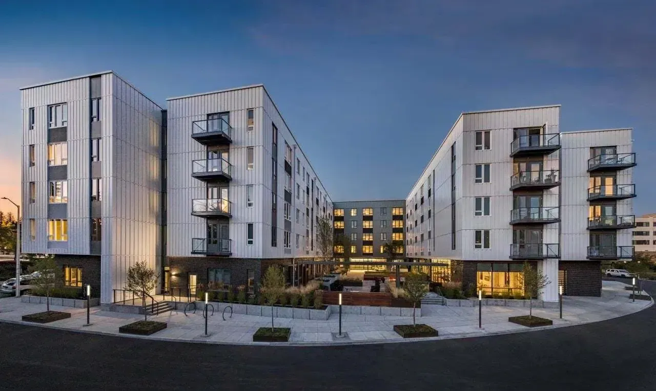A modern building with a white facade and balconies is surrounded by a landscaped area and a road.