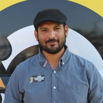 Man with a beard and newsboy cap smiles, wearing a company shirt in front of a logo.
