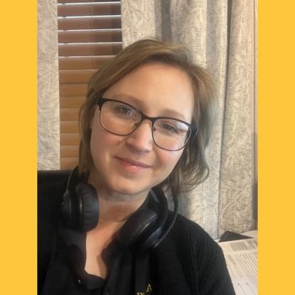 Woman with glasses and headphones smiling. Background: window blinds and curtain.