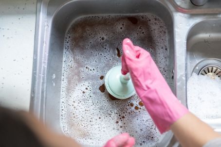 Person wearing pink gloves using a plunger in a dirty, soapy kitchen sink.