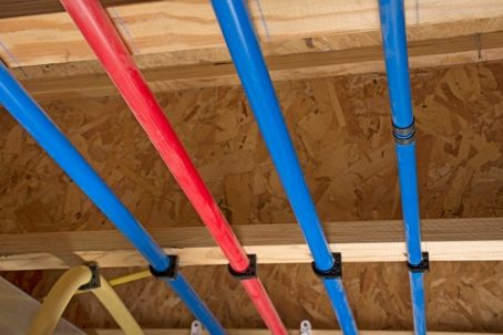 Blue and red PEX water pipes run across a wooden ceiling, held by black clamps.