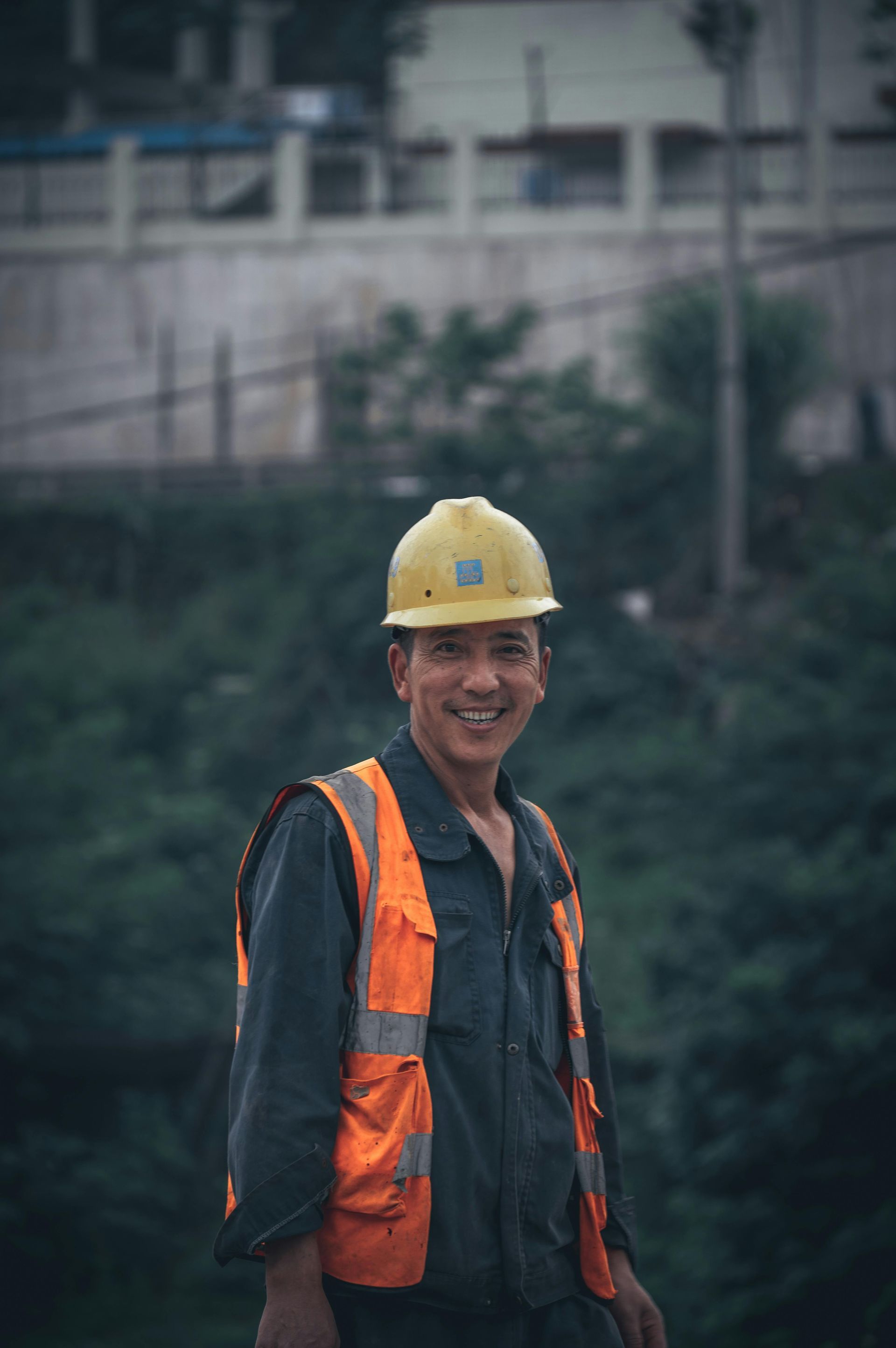 Construction worker in yellow hard hat and orange safety vest standing outdoors, smiling at the camera.