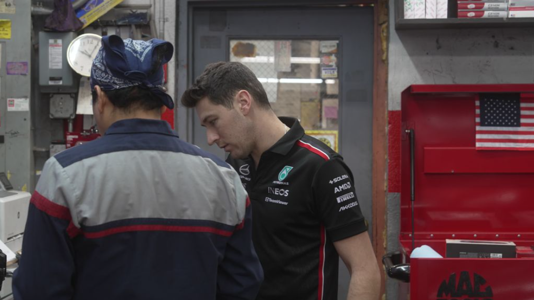 Two men in team uniforms talking in a workshop, with red tool cabinets in the background.