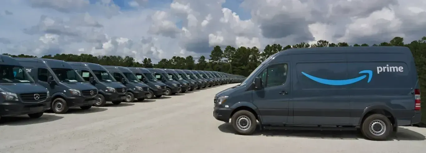 Rows of Amazon Prime delivery vans parked along a lot under a partly cloudy sky.