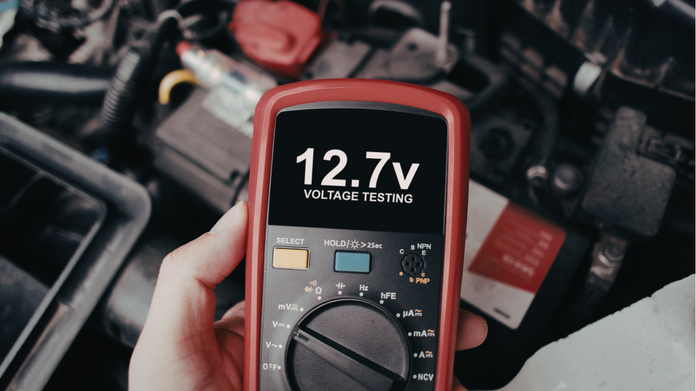 Hand holding a red digital multimeter showing 12.7V amid a cluttered electronics workbench
