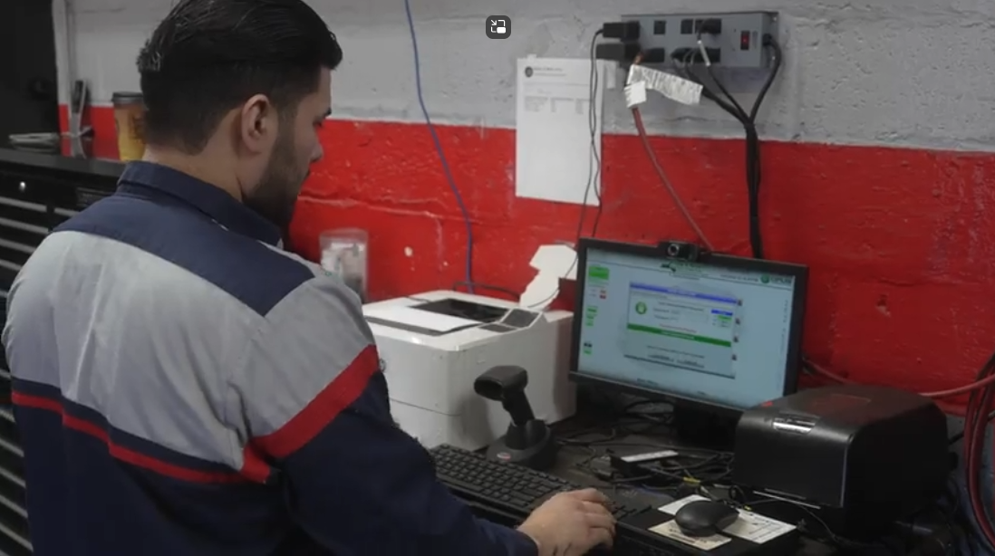 Worker in blue and gray jacket using a computer at a desk in an industrial workshop.