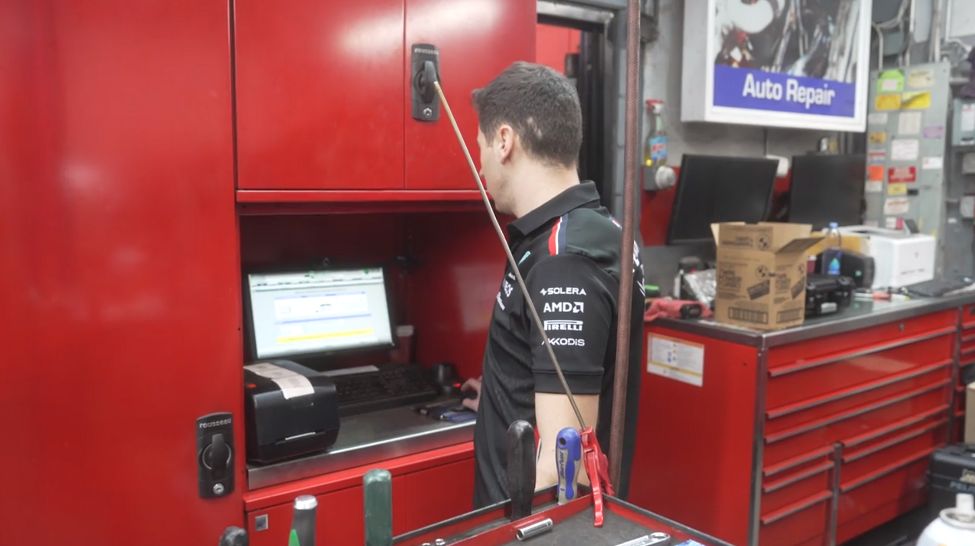 Mechanic working at a computer in an auto repair shop with red tool cabinets and machinery around him