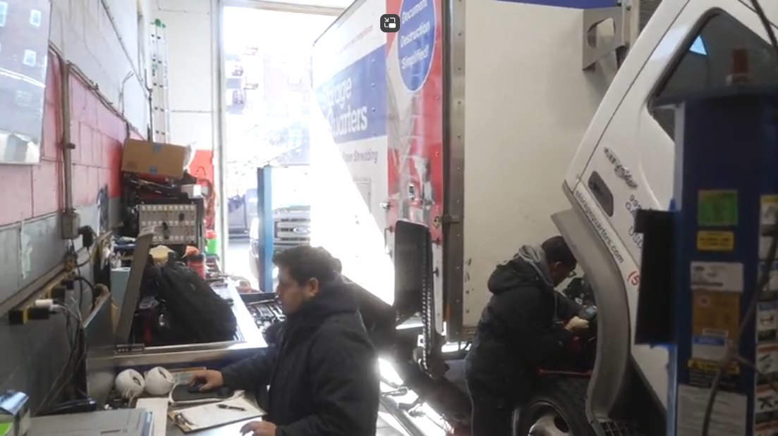 People working inside a narrow auto repair garage with a car raised on a lift.