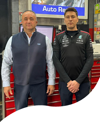 Two men in team jackets standing in a workshop with auto repair equipment and a red tool chest.