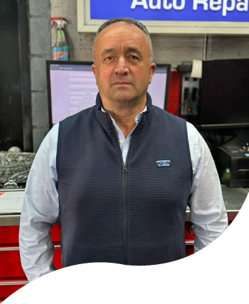 Man standing in an auto repair shop, wearing a navy vest and light blue shirt.
