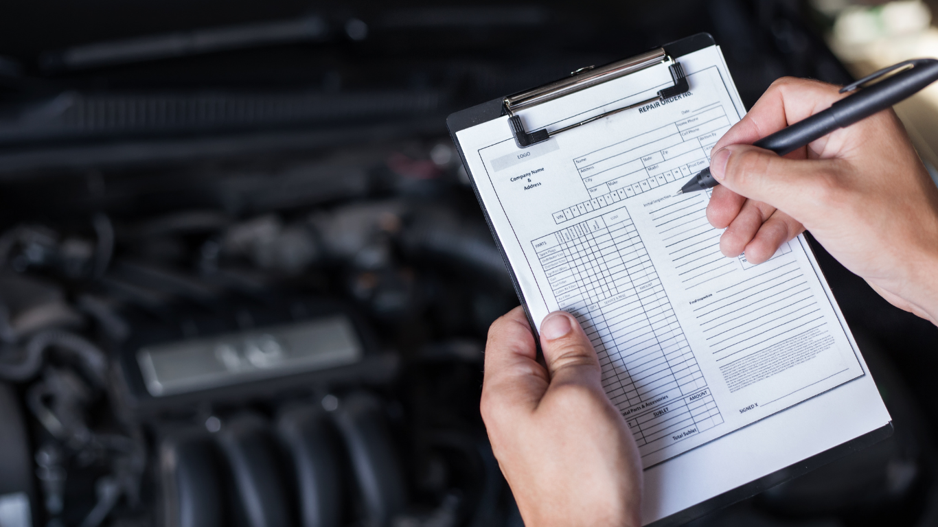 Hands using a clipboard and pen to inspect a car engine in a garage.