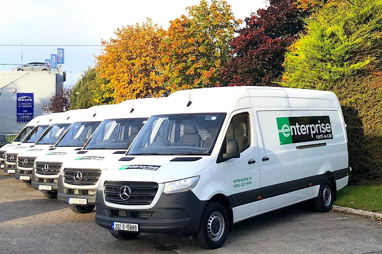 Row of white Enterprise rental vans parked outdoors beside autumn trees