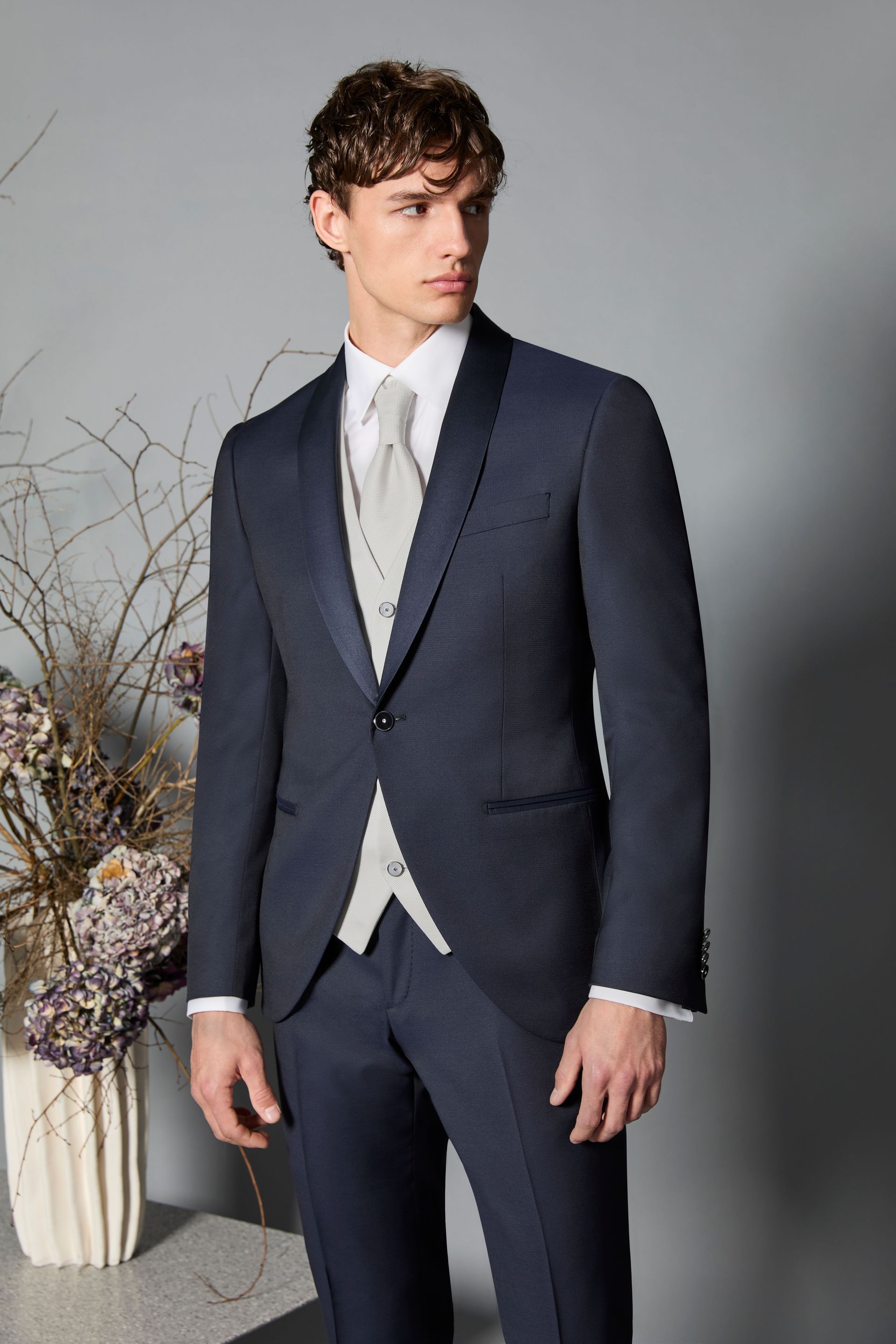 Uomo in abito blu navy con risvolto nero, camicia bianca, cravatta chiara e gilet, in posa in un interno.