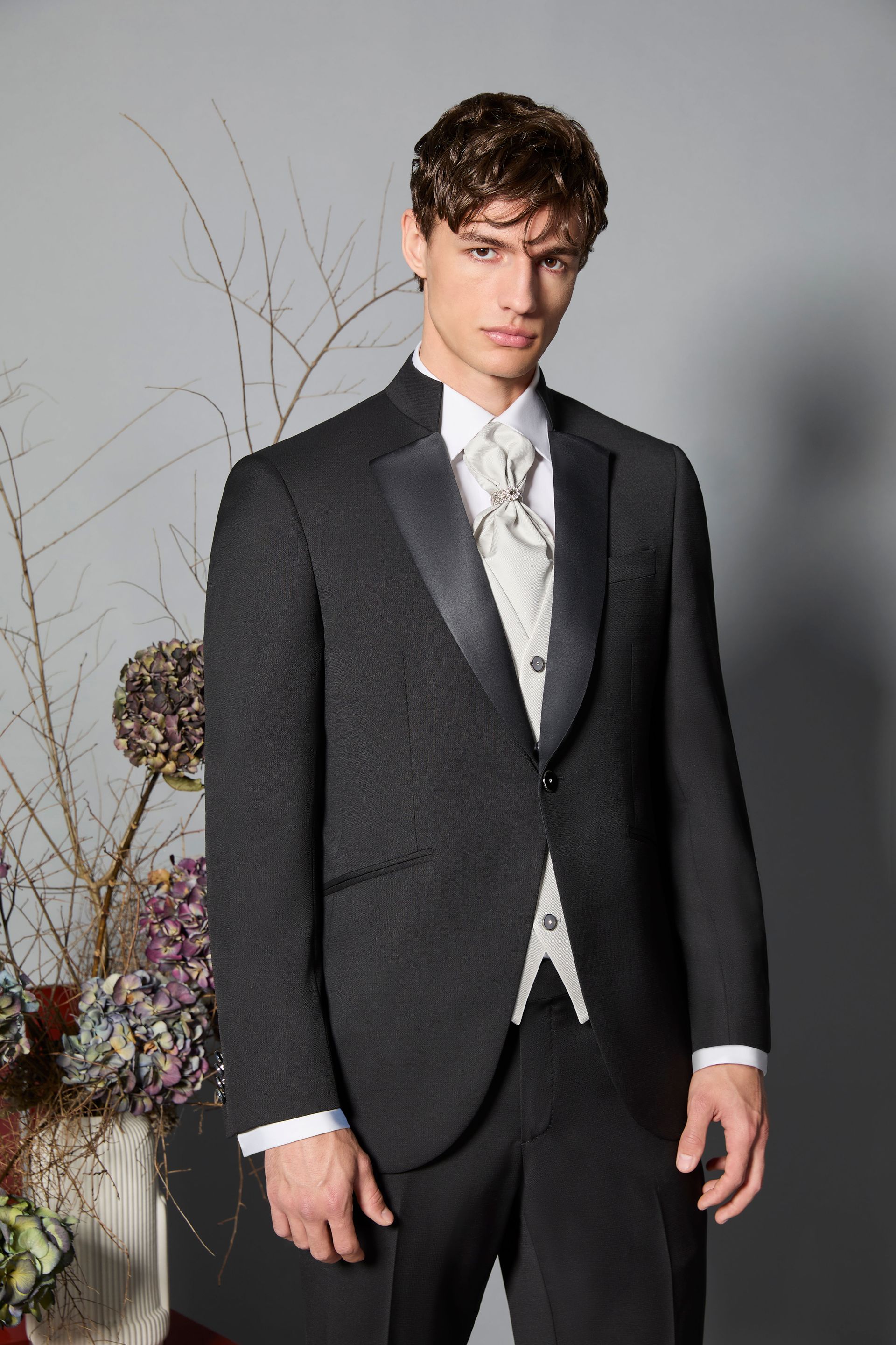 Uomo in abito nero formale con gilet bianco, in piedi davanti a decorazioni floreali.