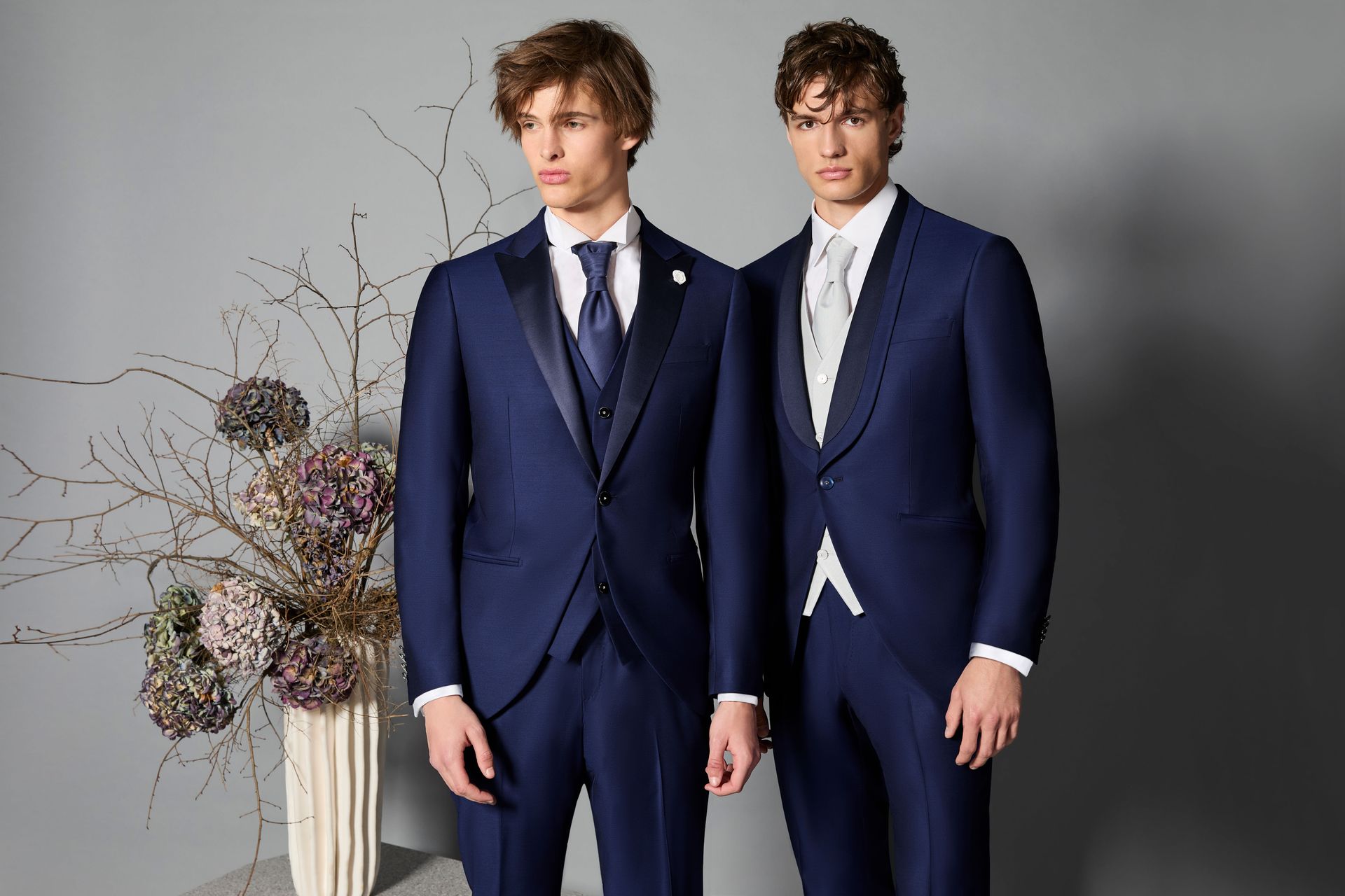 Due uomini in abiti blu navy con boutonniere, in piedi davanti a uno sfondo grigio.