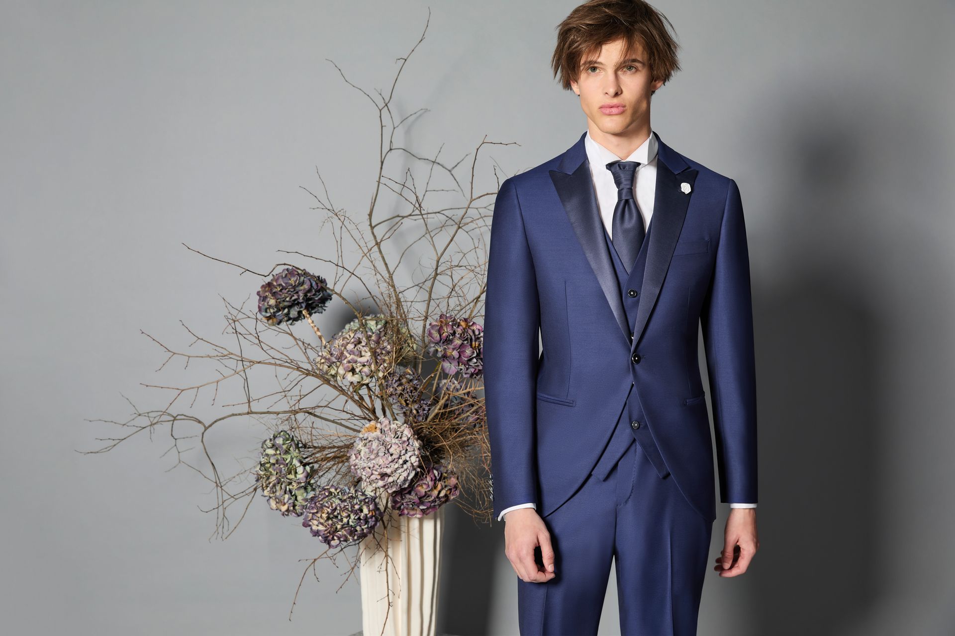 Uomo in abito blu navy con gilet e cravatta in piedi vicino a fiori secchi.