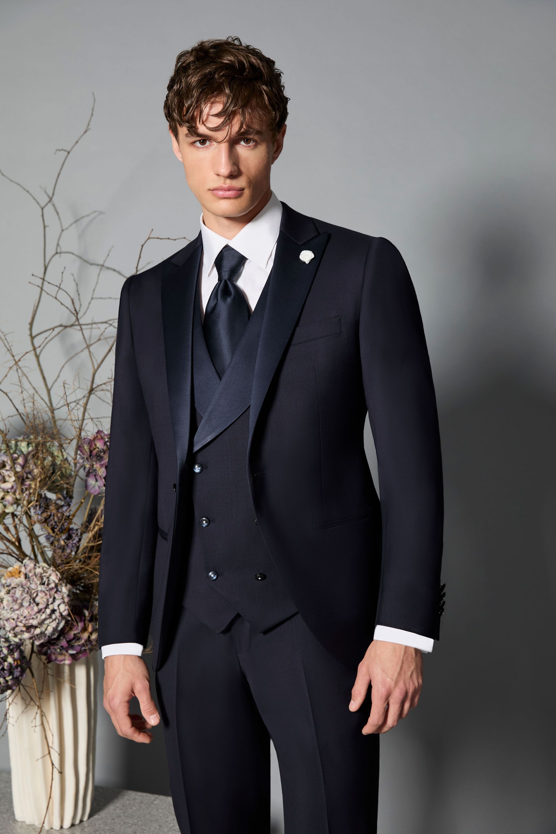 Uomo in abito blu navy, camicia bianca e cravatta, in posa formale in un interno.