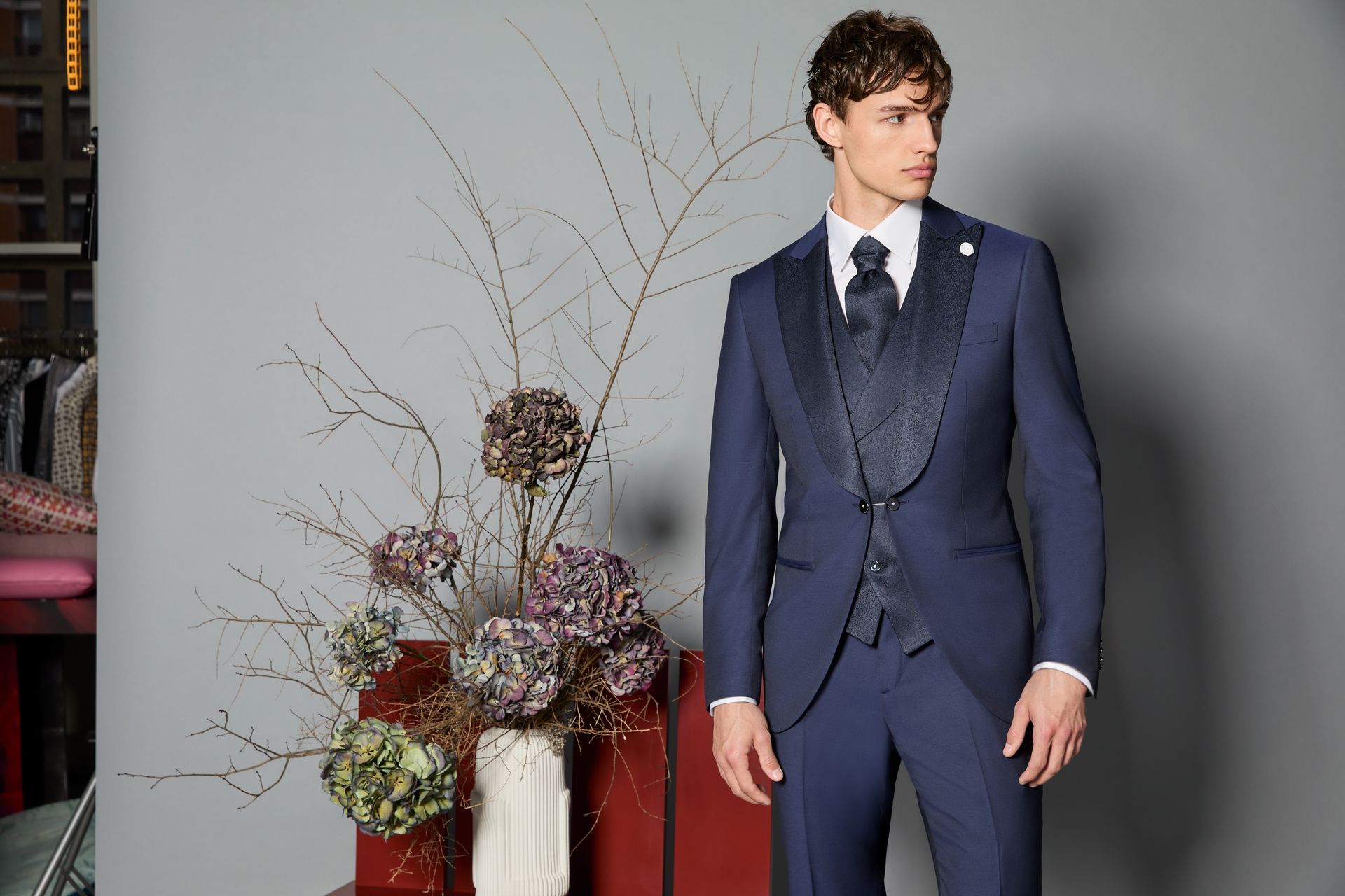 Uomo in abito blu navy, in piedi vicino a una composizione floreale. Abito scuro con risvolto fantasia. Sfondo grigio.
