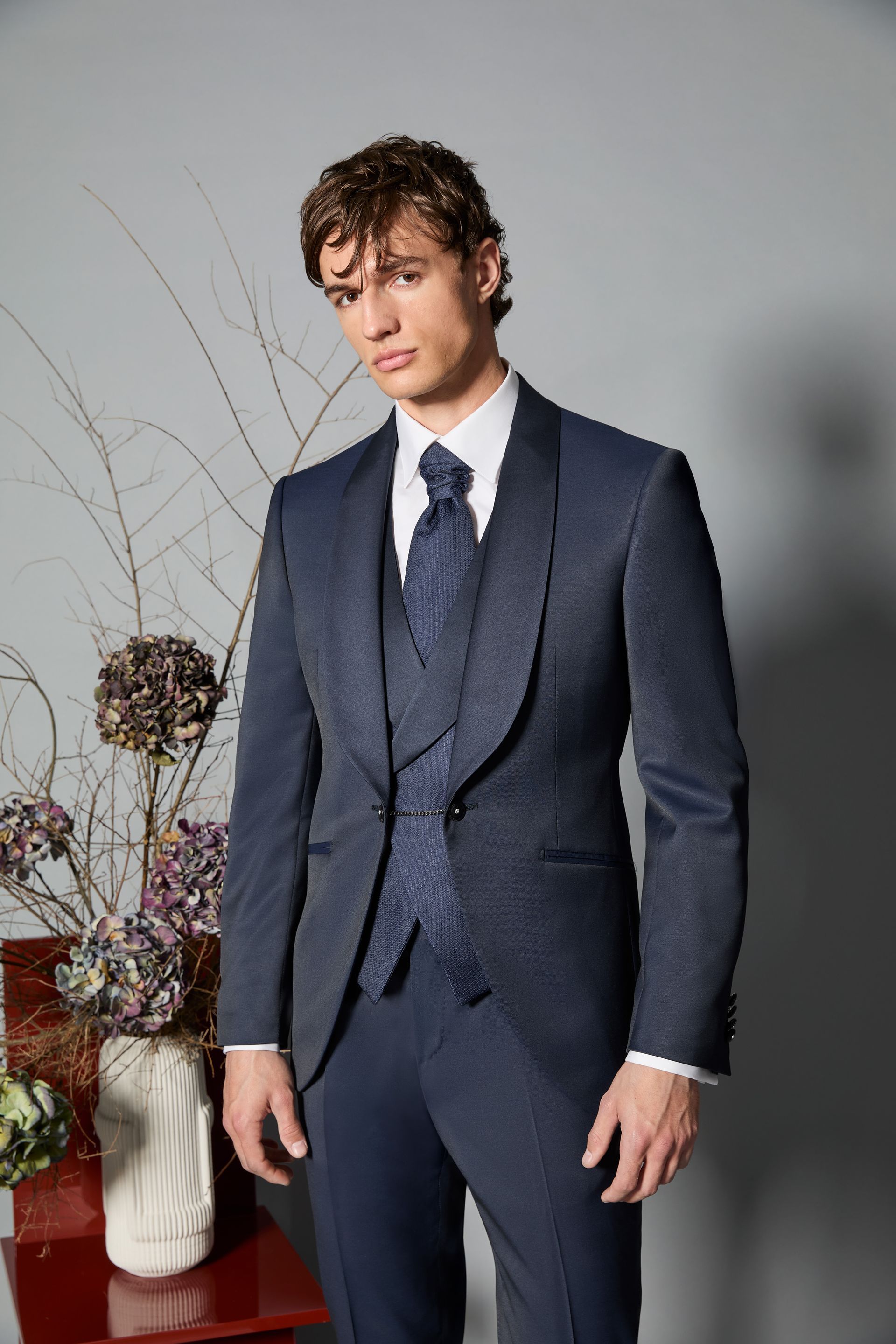 Uomo in smoking e gilet blu navy, camicia bianca e cravatta fantasia, in posa in un interno.