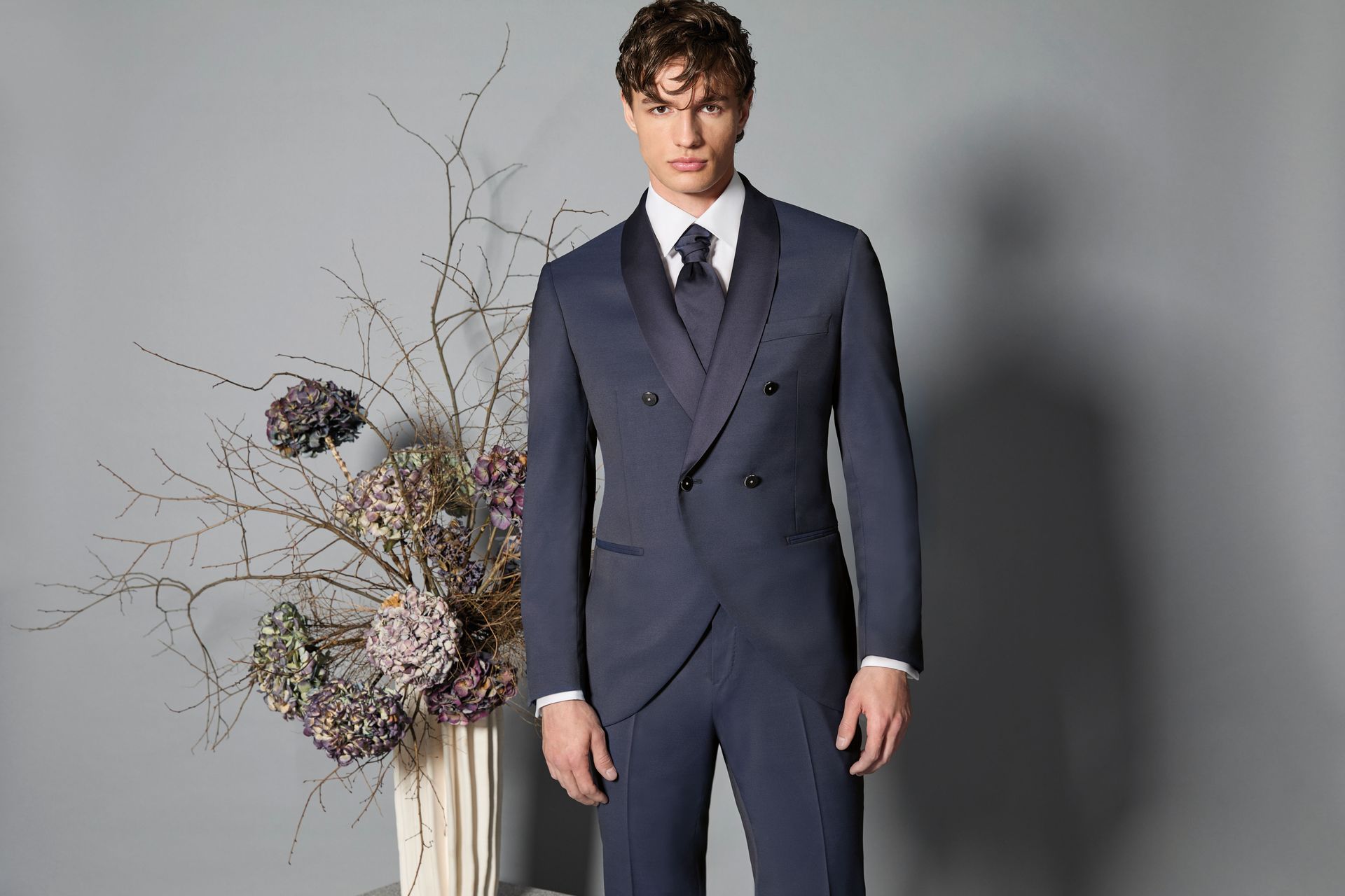 Uomo in abito blu, in piedi vicino a un vaso di fiori. Camicia bianca e cravatta. Sfondo grigio.