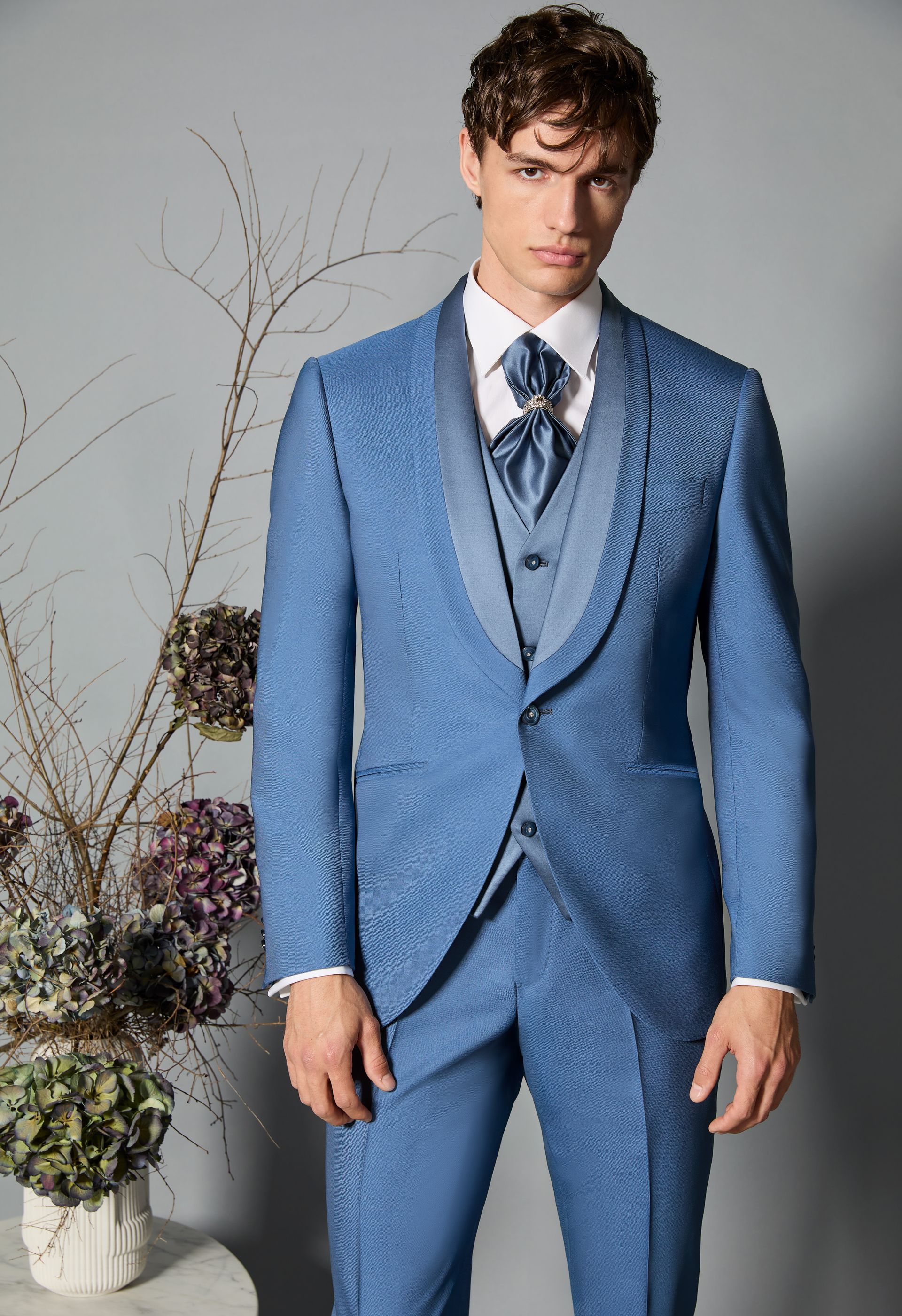 Uomo in abito blu con gilet e cravatta, in piedi davanti a uno sfondo neutro con decorazioni floreali.