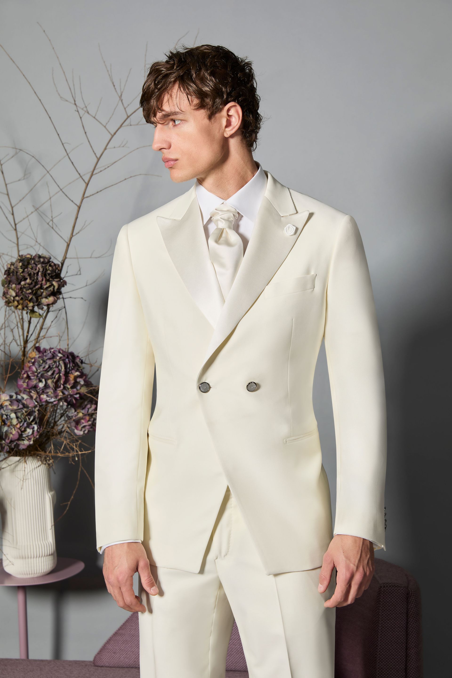 Uomo in abito doppiopetto color crema, camicia bianca e cravatta, in piedi in un interno con uno sfondo neutro.