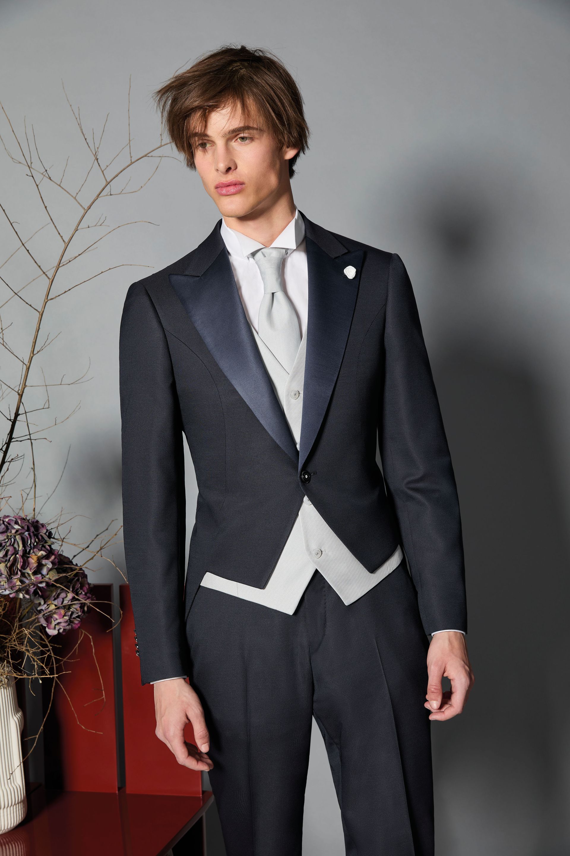 Uomo in smoking blu navy con gilet bianco e cravatta fantasia, in piedi in uno studio fotografico.