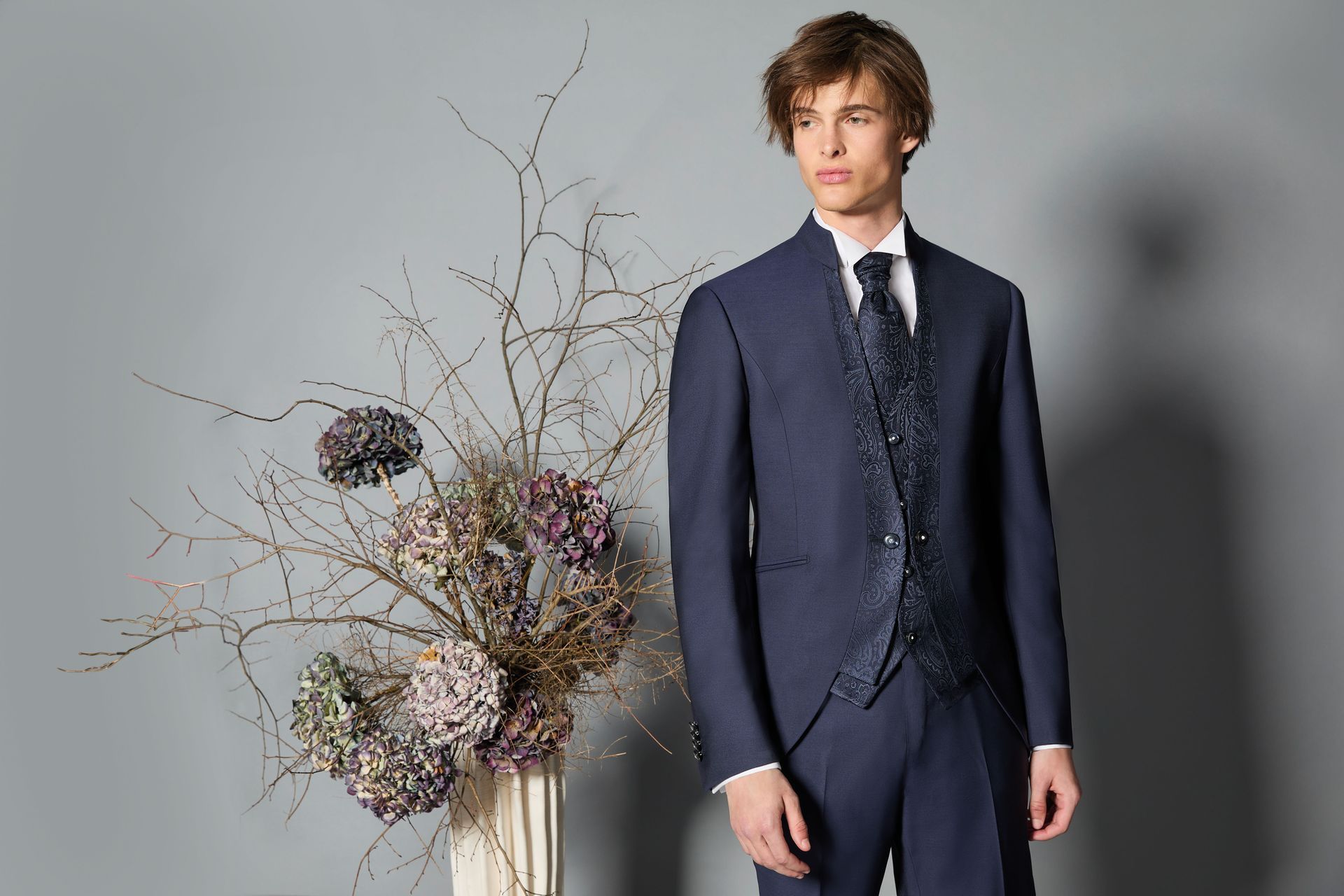 Uomo in abito blu navy, in piedi vicino a un vaso con composizione floreale; sfondo grigio.