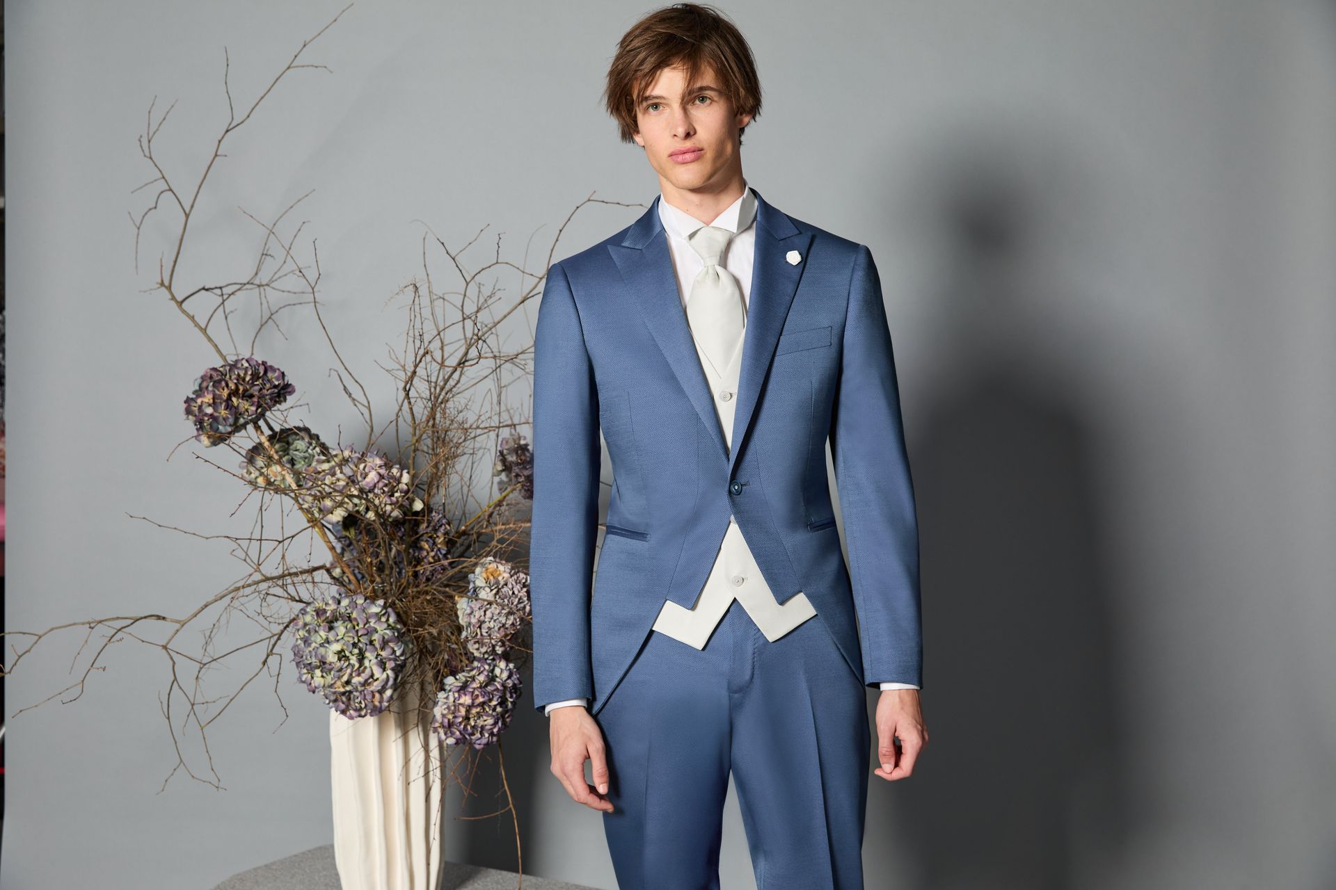Uomo in abito blu e gilet con camicia bianca e cravatta, in posa vicino ai fiori.