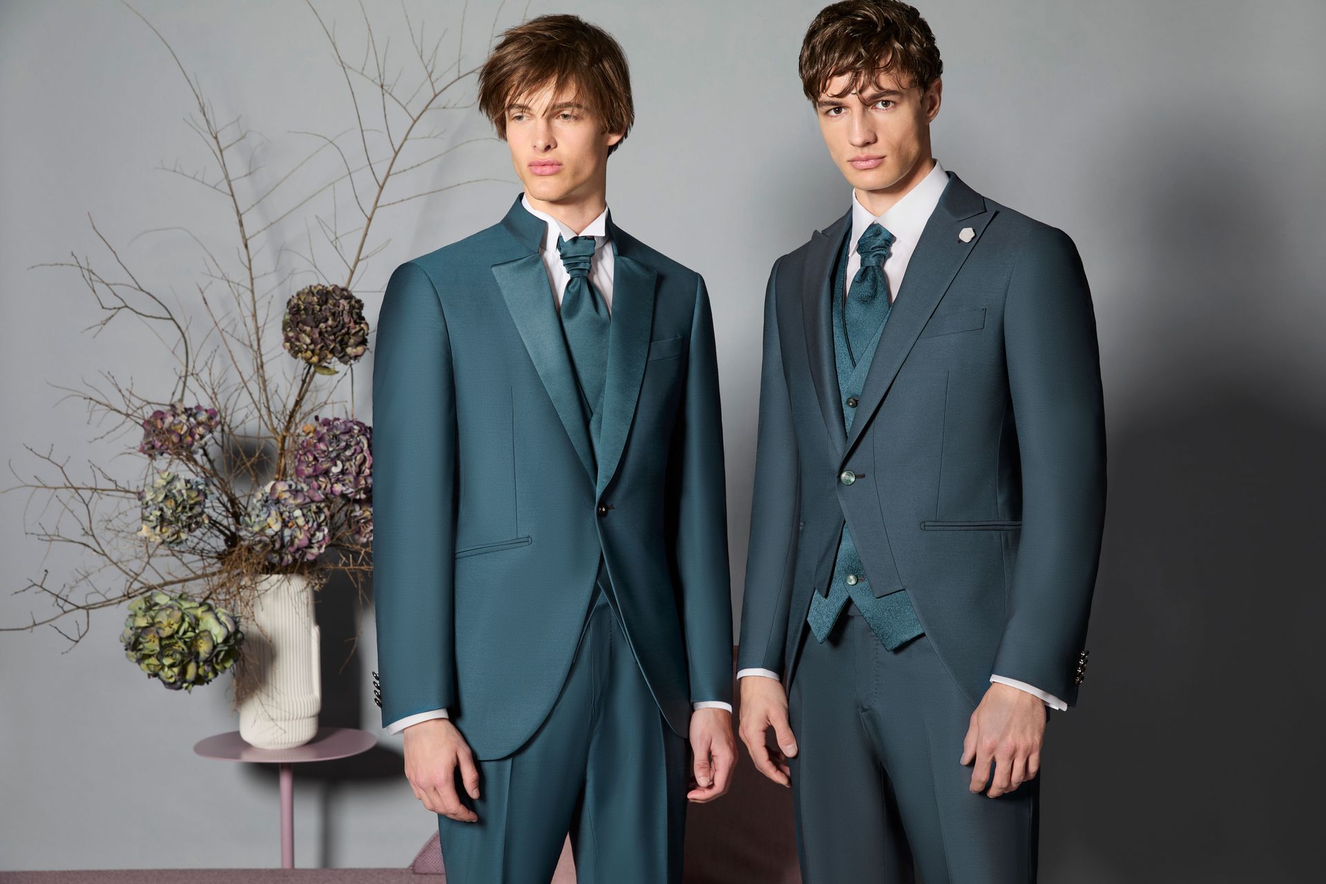 Due uomini in abiti color verde acqua posano accanto a una composizione floreale; uno degli uomini indossa un gilet.