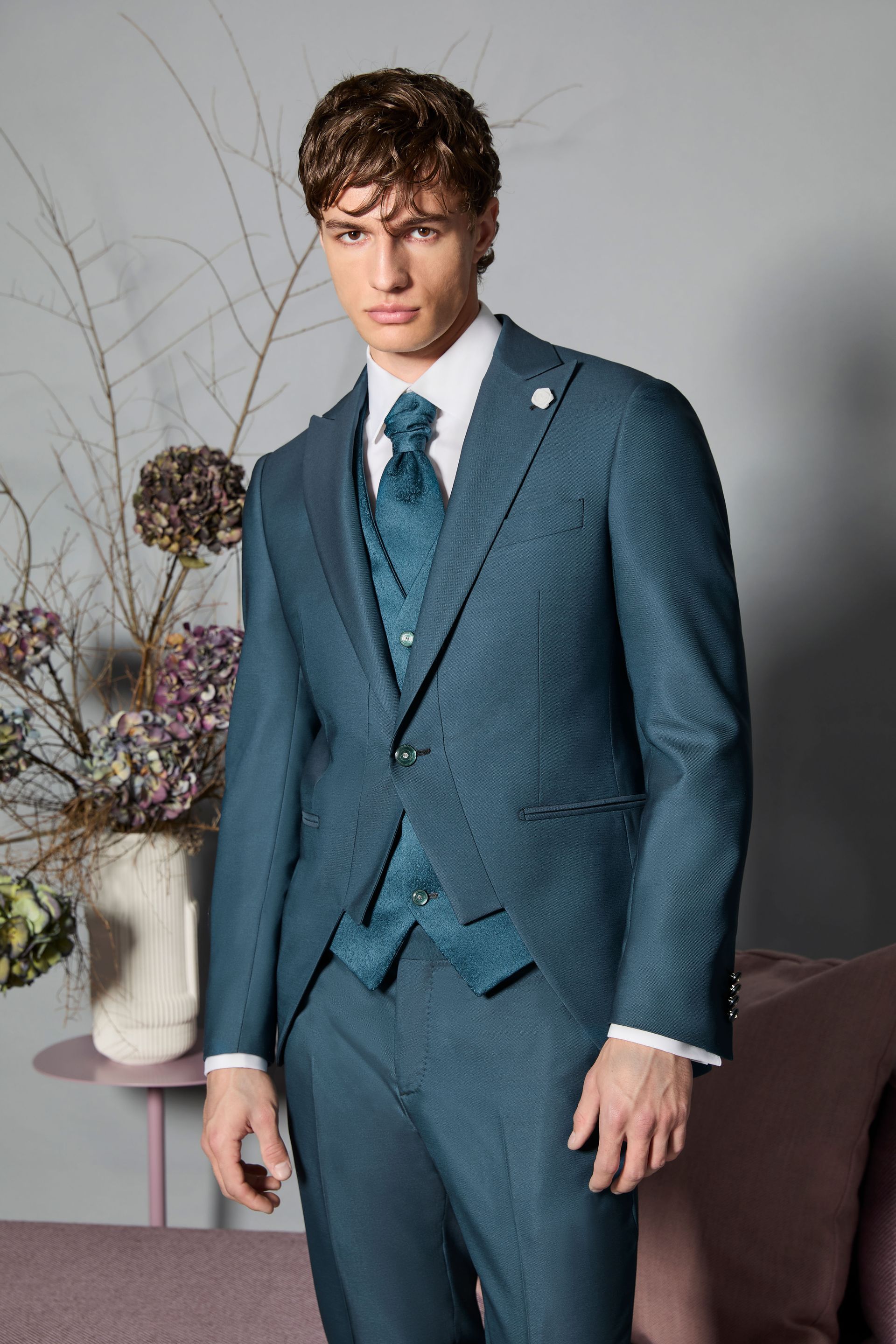 Uomo in abito e gilet verde acqua, camicia bianca, spilla floreale, in posa in un interno.