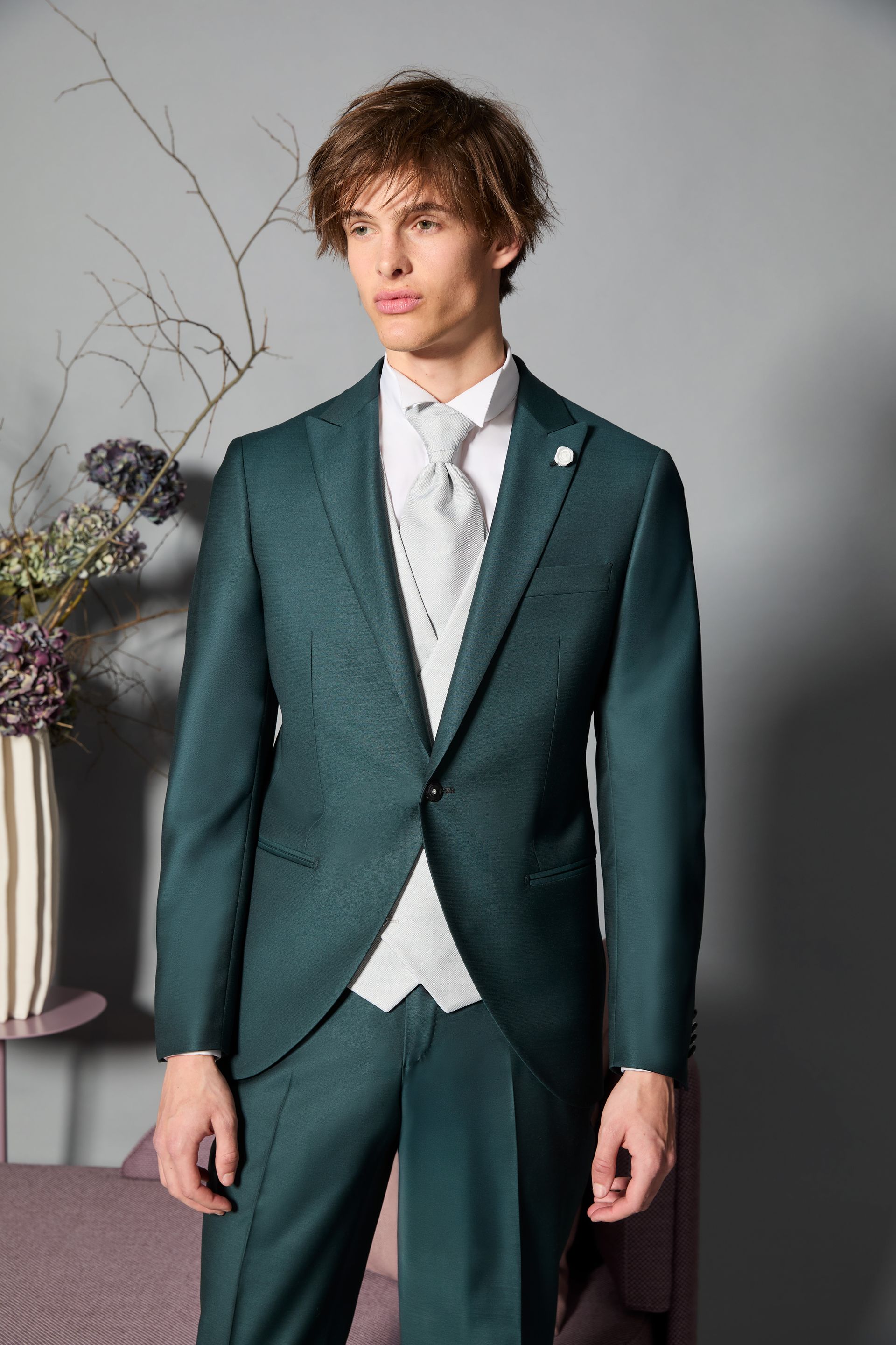 Uomo in abito verde acqua e gilet, abbottonato, che guarda di lato. Ambiente neutro.