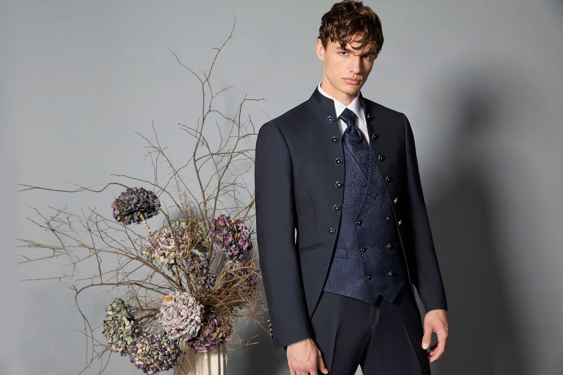 Uomo in abito blu navy e gilet fantasia, in piedi accanto a una composizione floreale su sfondo grigio.