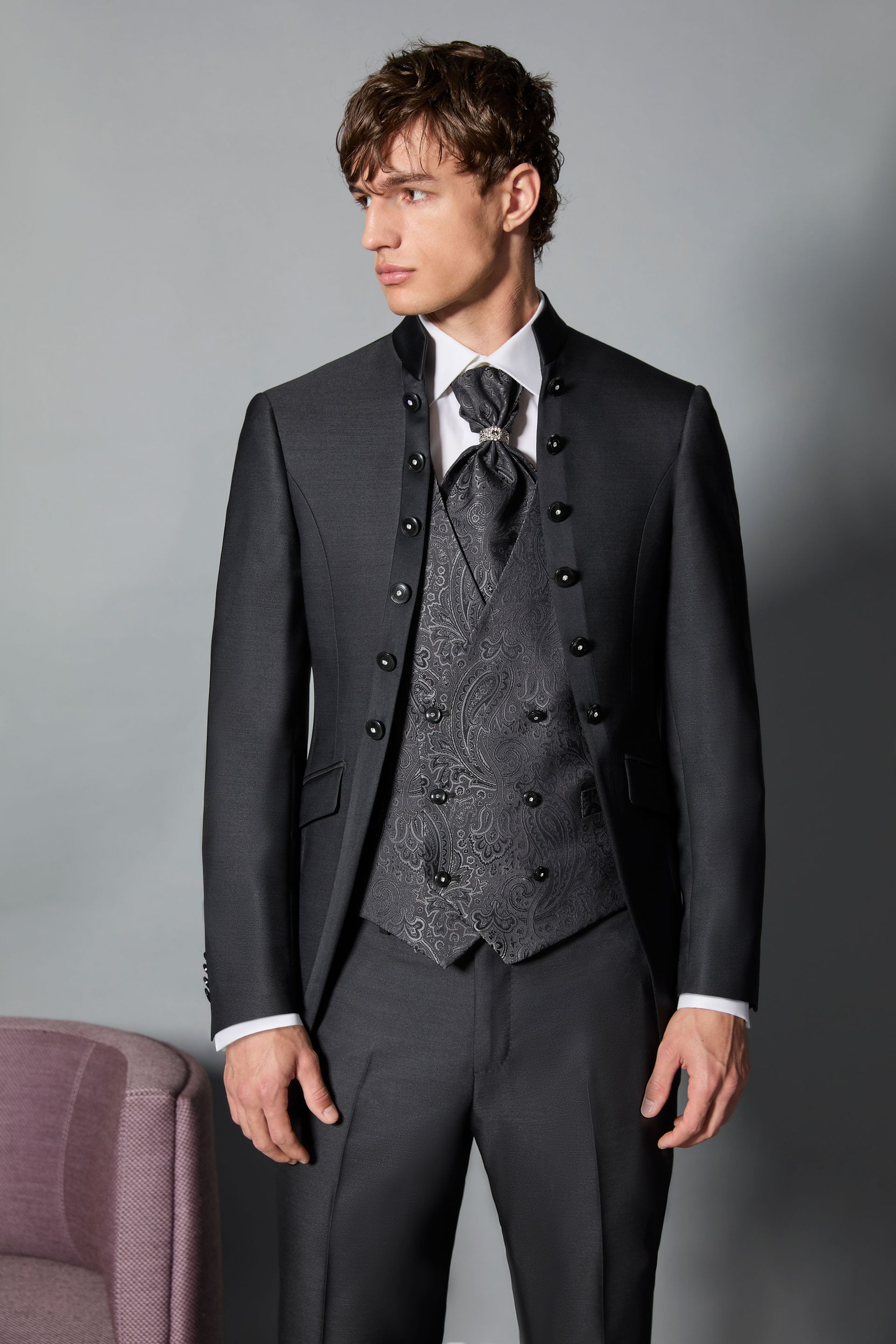 Uomo in abito grigio scuro con gilet, in piedi su uno sfondo grigio.