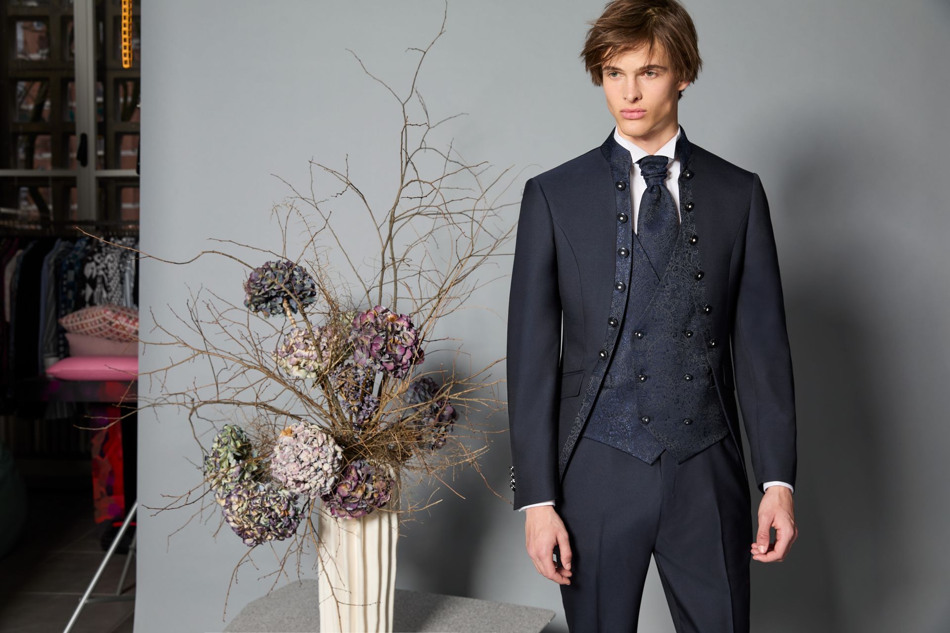 Un uomo in abito blu navy con gilet decorato è in piedi vicino a una composizione floreale in studio.