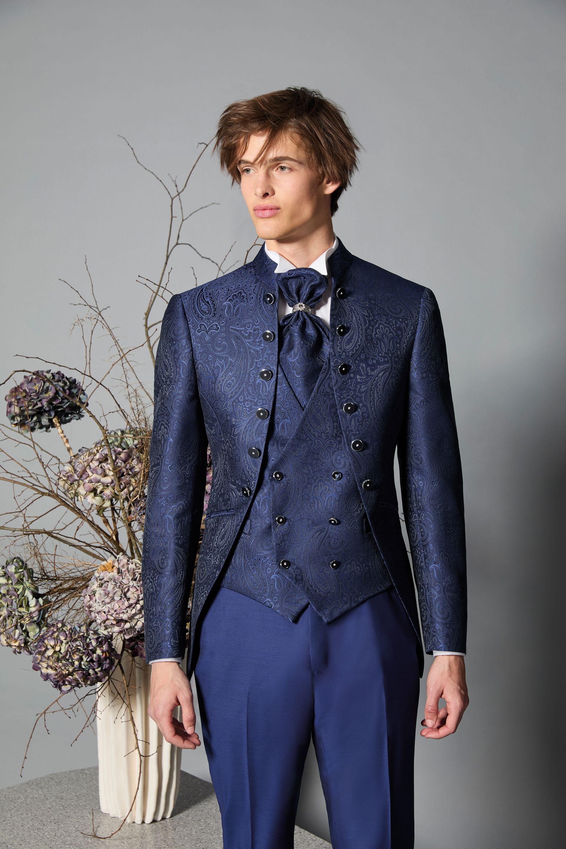 Uomo in un elegante abito blu con giacca a fantasia, gilet e cravatta, in piedi in un ambiente interno.